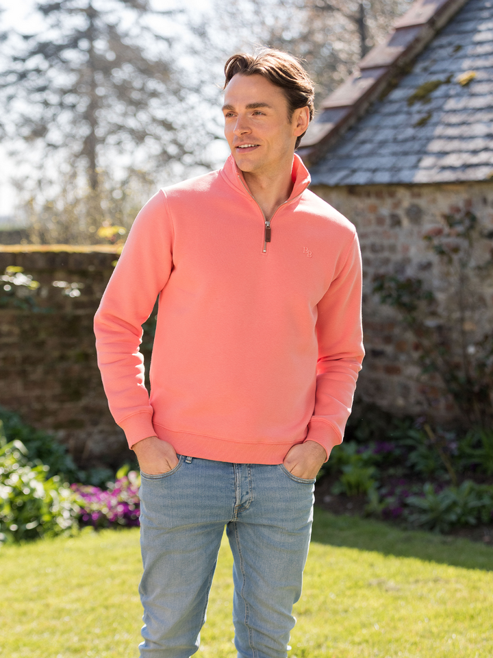 Man wearing a flamingo (coral) pink quarter-zip pullover sweatshirt with subtle embroidered logo, styled casually with light blue jeans.