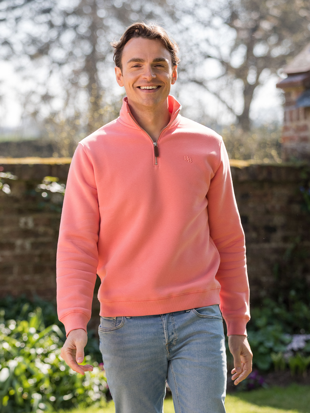 Man wearing a flamingo (coral) pink quarter-zip pullover sweatshirt with subtle embroidered logo, styled casually with light blue jeans.