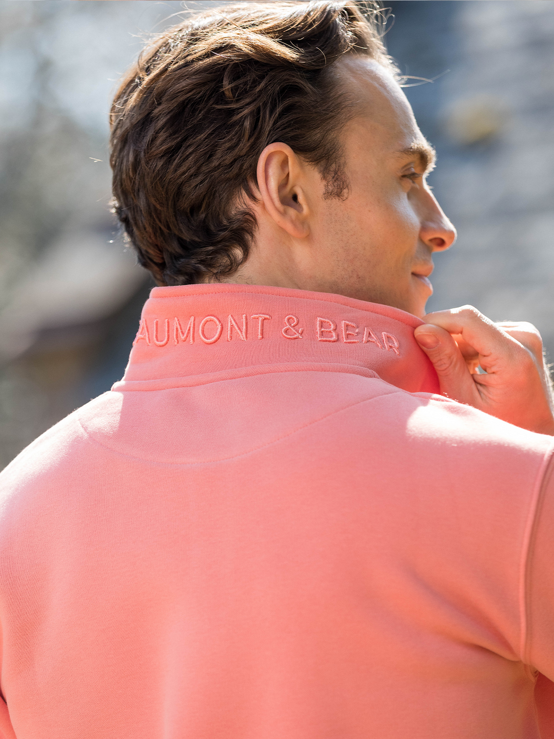 Man wearing a flamingo (coral) pink quarter-zip pullover sweatshirt with subtle embroidered logo, styled casually with light blue jeans.