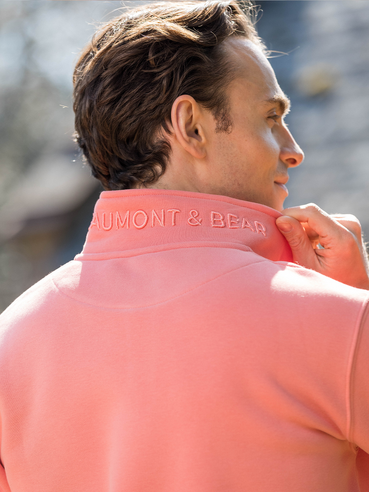Man wearing a flamingo (coral) pink quarter-zip pullover sweatshirt with subtle embroidered logo, styled casually with light blue jeans.