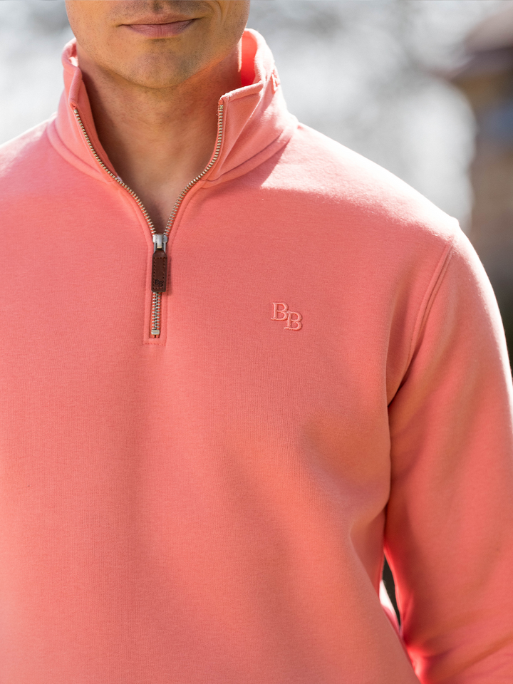 Man wearing a flamingo (coral) pink quarter-zip pullover sweatshirt with subtle embroidered logo, styled casually with light blue jeans.