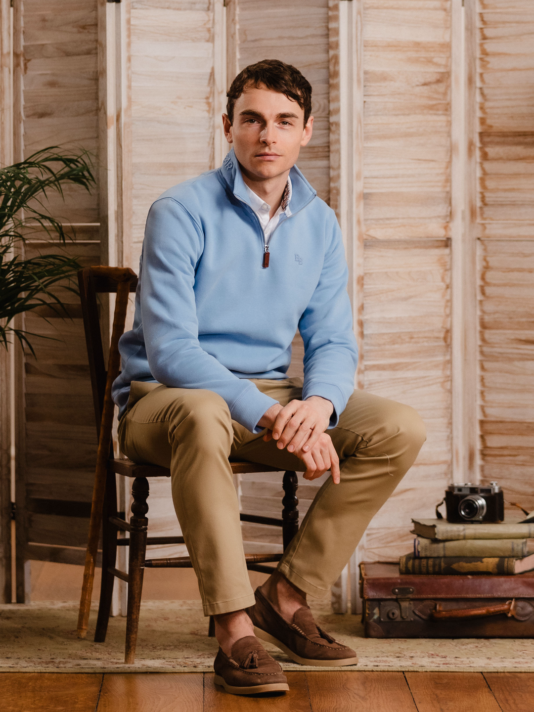 Man wearing a cornflower blue (light blue) quarter-zip pullover sweatshirt with subtle embroidered logo, styled casually with tan trousers. 