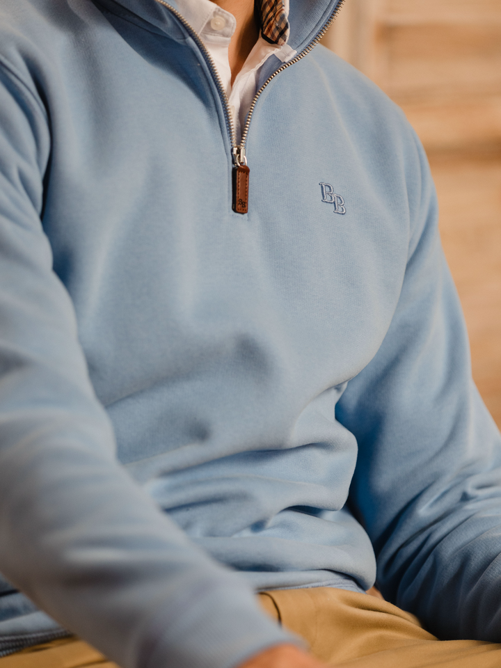 Man wearing a cornflower blue (light blue) quarter-zip pullover sweatshirt with subtle embroidered logo, styled casually with tan trousers. 
