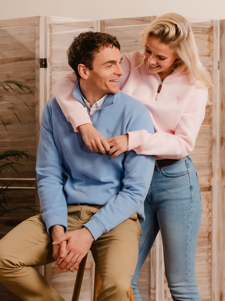 Man wearing a cornflower blue (light blue) quarter-zip pullover sweatshirt with subtle embroidered logo, styled casually with tan trousers. 