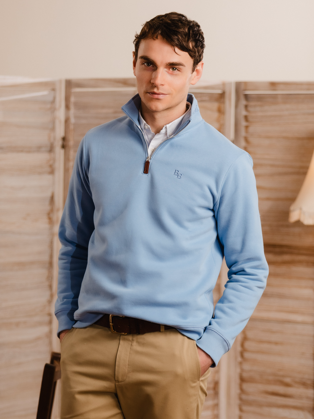Man wearing a cornflower blue (light blue) quarter-zip pullover sweatshirt with subtle embroidered logo, styled casually with tan trousers. 