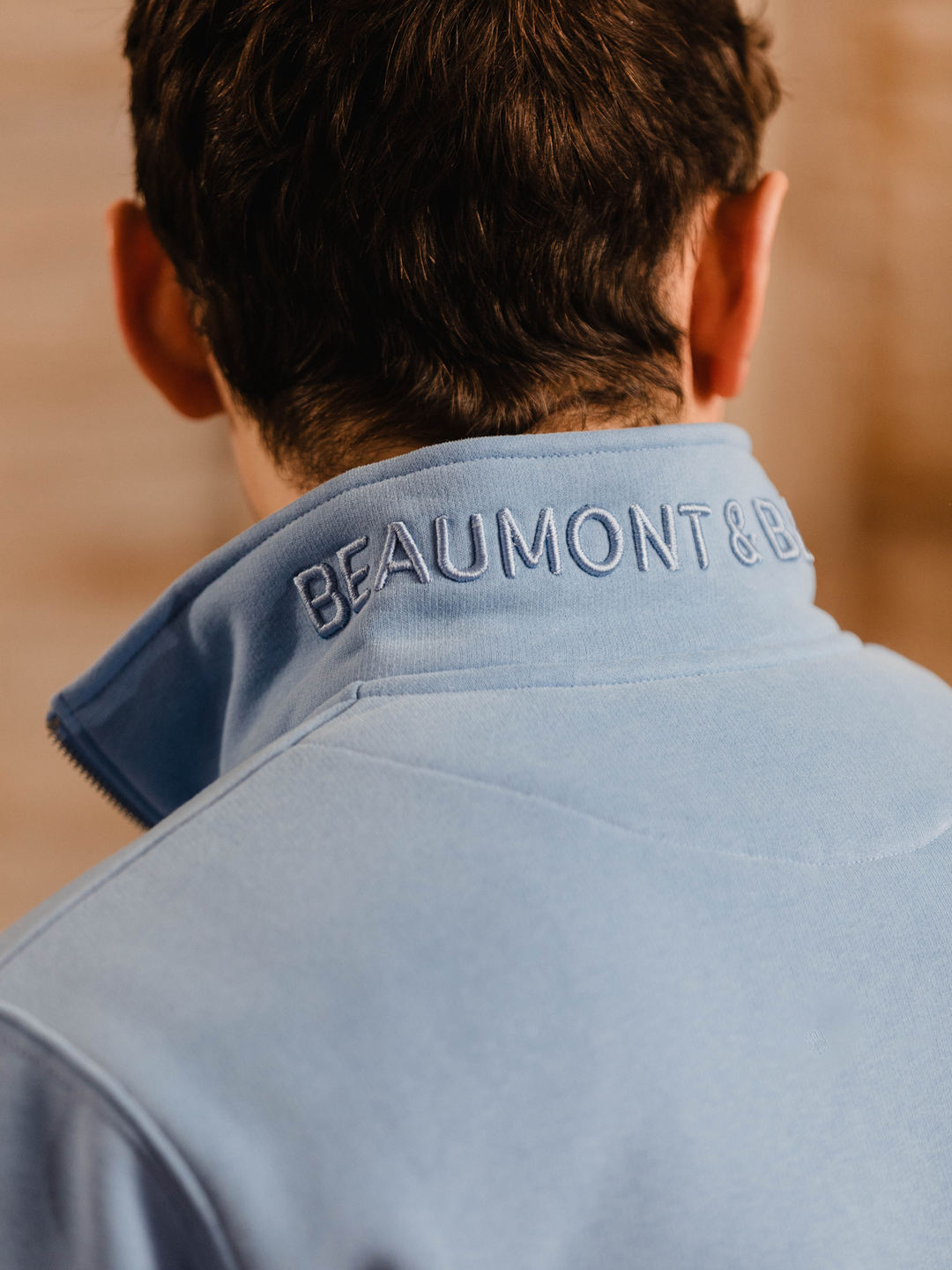 Man wearing a cornflower blue (light blue) quarter-zip pullover sweatshirt with subtle embroidered logo, styled casually with tan trousers. 