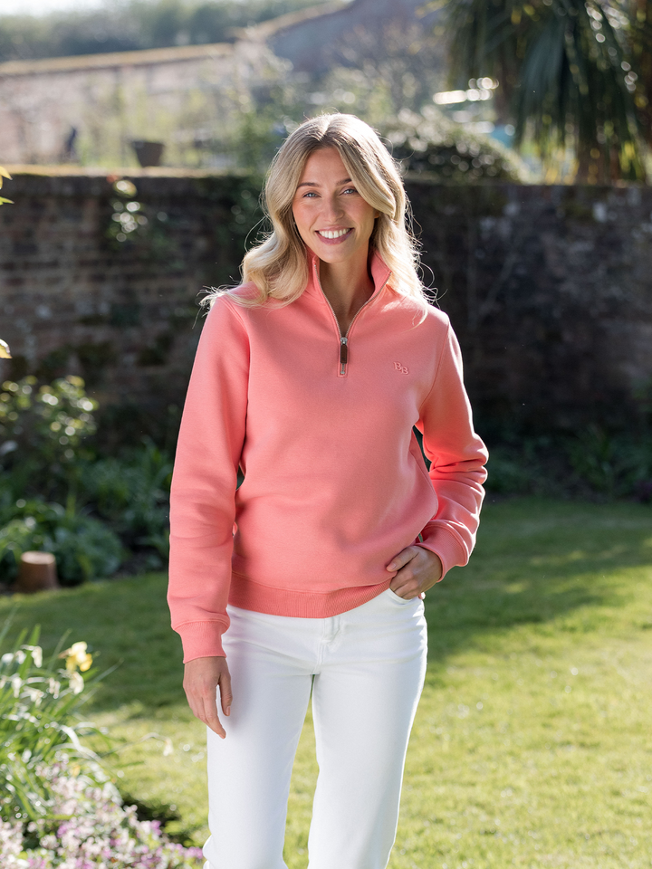 Woman wearing a flamingo (coral) pink quarter-zip pullover sweatshirt with subtle embroidered logo, styled casually with white jeans. 