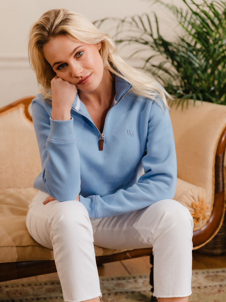 Women wearing a cornflower blue (light blue) quarter-zip pullover sweatshirt with subtle embroidered logo, styled casually with white jeans.